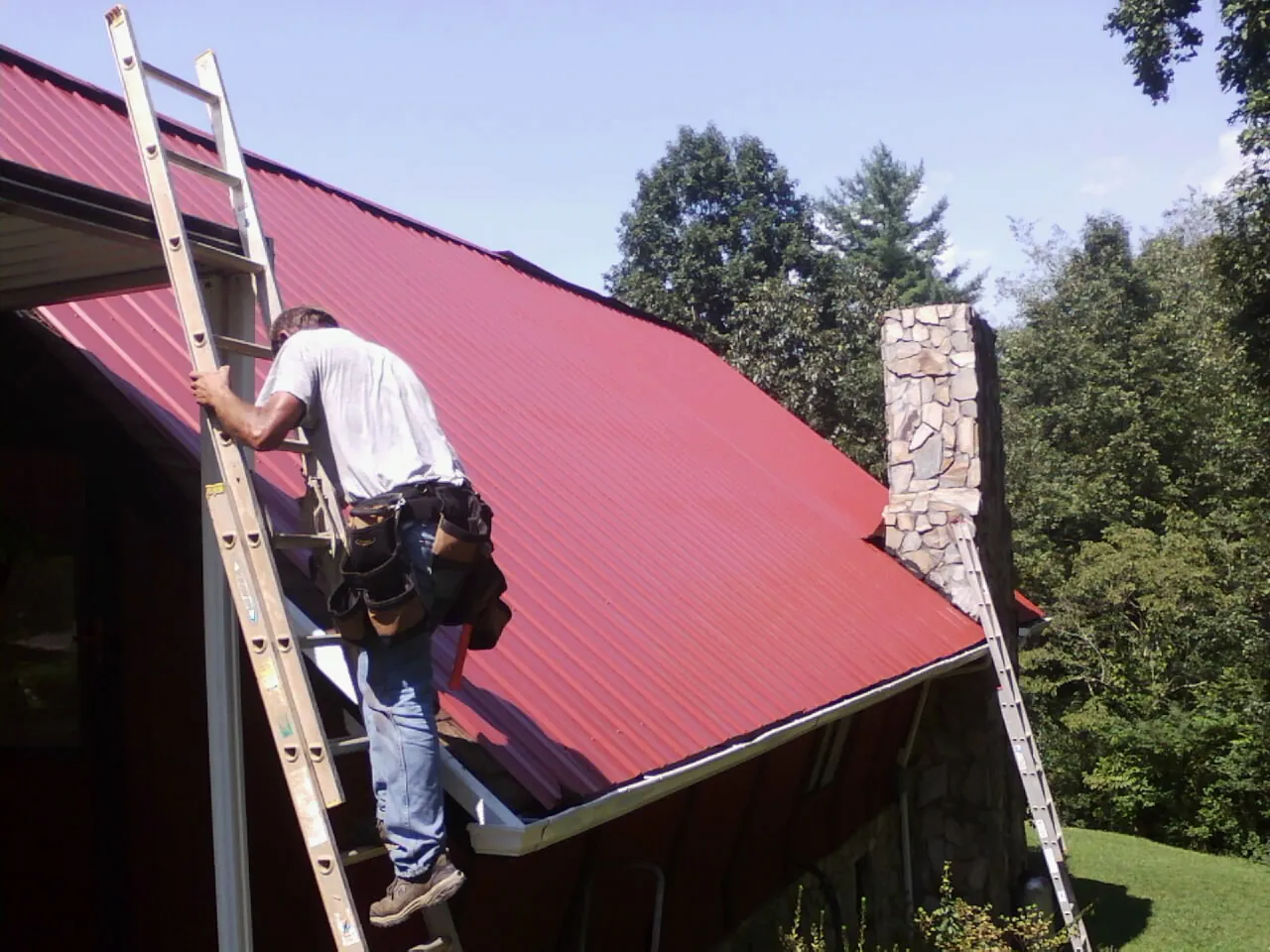 Roofer on a ladder beside a red metal roof and stone chimney — Soffit & Fascia Repair by Roofing Brothers in Watonga
